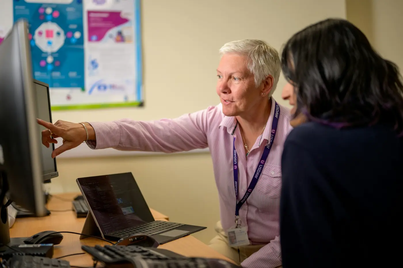 A worker with a badge pointing at a computer screen while helping a colleague